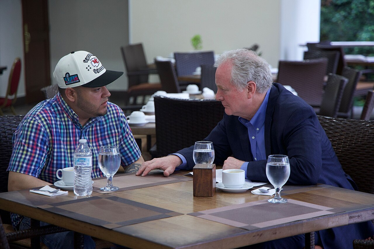 Kilmar Abrego Garcia, wearing a white baseball cap and plaid shirt, seated at a restaurant table speaking with Senator Chris Van Hollen in El Salvador